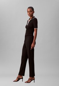 Polo nera in maglia con dettaglio del colletto bianco, abbinata a pantaloni neri su misura a vita alta e tacchi neri con cinturini su sfondo grigio.