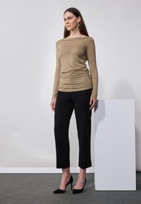 Beige langermet topp med samlet detalj, kombinert med svarte ankel-lange bukser og svarte høye hæler, satt mot en nøytral bakgrunn.