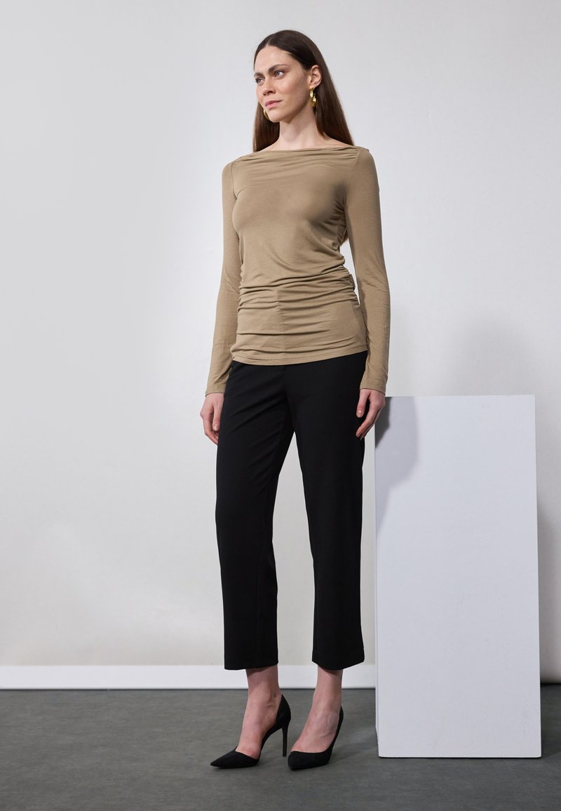 Beige langermet topp med samlet detalj, kombinert med svarte ankel-lange bukser og svarte høye hæler, satt mot en nøytral bakgrunn.