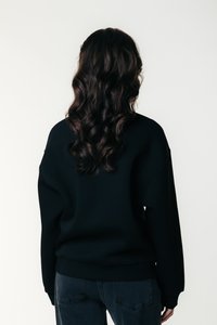 Black oversized sweatshirt with a rounded neckline, long sleeves, and ribbed cuffs. Worn with dark denim jeans, featuring a textured back view.