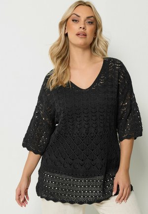 Blonde vrouw draagt een zwart gehaakt topje met ellelange mouwen en een geschulpte zoom, gecombineerd met een witte broek.