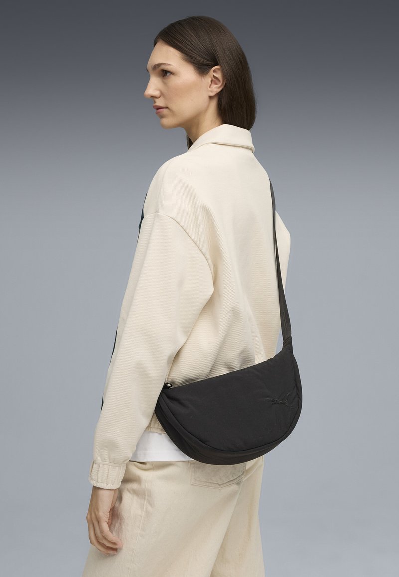 Czarna torebka typu crossbody o zakrzywionym kształcie i regulowanym pasku, noszona na biodrze na tle jasnobeżowej bluzki i spodni. Minimalistyczny design.
