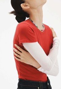 Camiseta roja de manga corta sobre una blusa blanca de manga larga, combinada con jeans de mezclilla oscura. El atuendo incluye un collar de cuentas como detalle destacado.