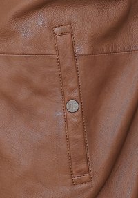 Braunes Lederprodukt mit einem auffälligen Druckknopf und genähten Akzenten. Die Oberfläche hat eine glatte Textur mit dezenten Maserungsmustern.