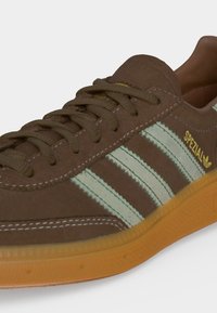 Καφέ σουέντ αθλητικό παπούτσι Adidas Spezial με λευκές ρίγες και λαστιχένια σόλα, με καφέ κορδόνια και κίτρινη σήμανση στο πλάι.