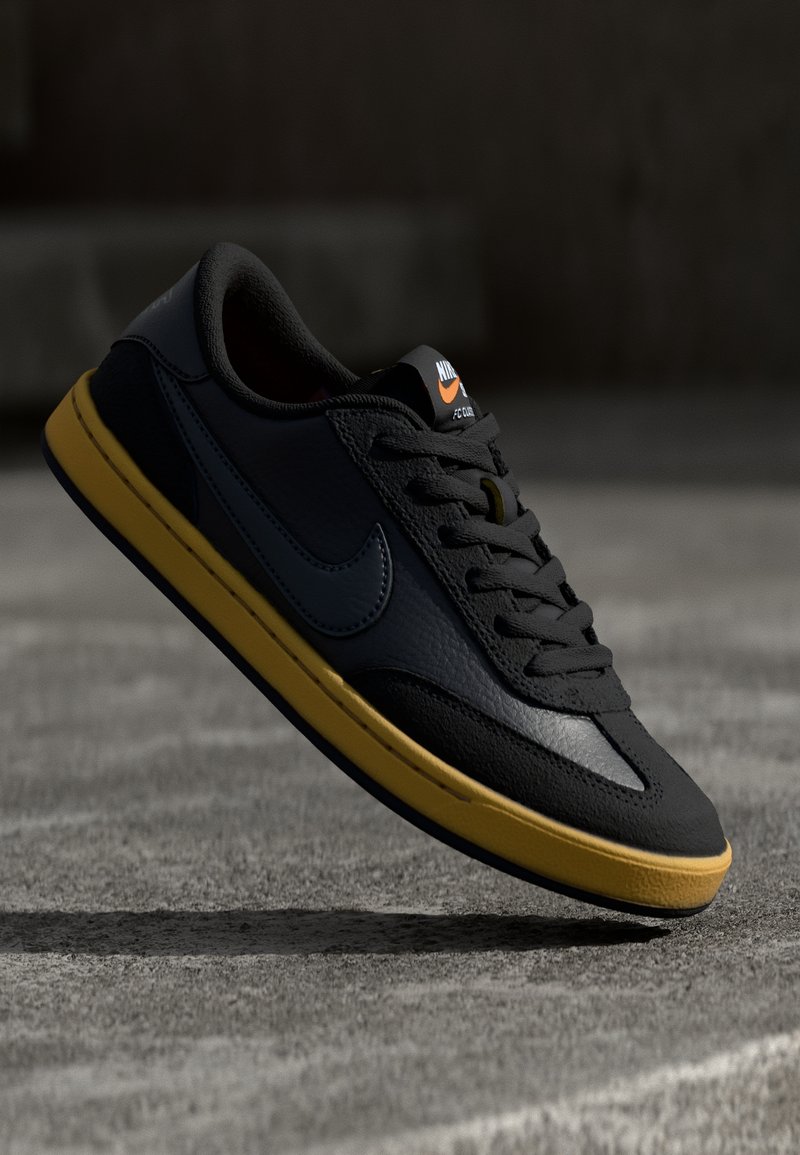 Musta nahkainen tennari, jossa on keltainen pohja, tasaiset nauhat, teksturoiduilla yksityiskohdilla ja näkyvä Nike-logo kielen ja sivun kohdalla.