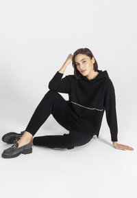 Czarna dziana bluza z kapturem oraz dopasowane spodnie, z poziomą srebrną prostą na piersi. Tkanina jest miękka i teksturowana. Uzupełniona o masywne czarne buty.