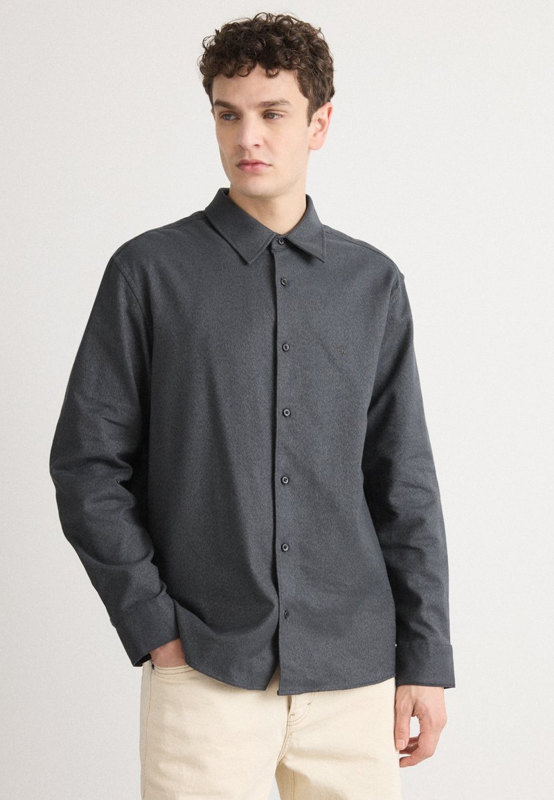 Camicia grigia scura a maniche lunghe con chiusura a bottoni, colletto a punta e una trama sottile. Include un dettaglio con il marchio sul petto.