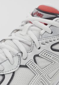 Bílé sportovní boty s vrchním dílem z prodyšné síťoviny, bílé šněrování a černé akcenty. Obsahují šedé detaily a červený polstrovaný límec. Povrch s texturovaným detailem.