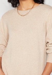Suéter de punto beige con cuello redondo y mangas largas. Presenta una textura suave y un corte holgado, complementado por un collar de cadena dorada.