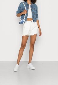 Woman wearing blue denim jacket, white crop top, white frayed shorts, white sneakers, and a small blue-and-white patterned shoulder bag.