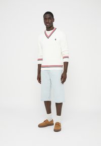 Baltas kabelio mezgimo megztinis su V formos iškirpte ir raudonomis bei juodomis juostomis. Dėvimas su šviesiai mėlynais džinsiniais šortais ir rudos spalvos zomšiniais batais.