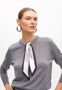 Szary sweter z okrągłym dekoltem i długimi rękawami, z dekoracyjnym kokardowym paskiem w białym i czarnym kolorze z napisami. Duże srebrne kolczyki w kształcie obręczy.