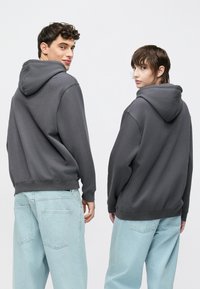 Szürke kapucnis pulóverek kenguruzsebbel, bordázott mandzsettával és szegéllyel, világoskék bő nadrággal viselve, hátulnézetből látható.