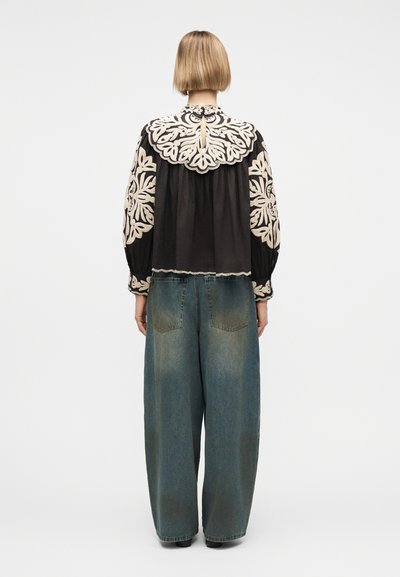 Personne aux cheveux blonds courts portant un chemisier noir avec de grandes broderies florales crème et un jean bleu délavé ample, debout devant un fond blanc.