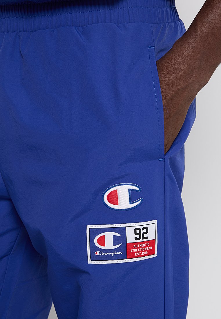 Main droite dans la poche de shorts de sport Champion bleus arborant des logos Champion et un patch avec le texte "92 Authentic Athleticwear Est.1919".