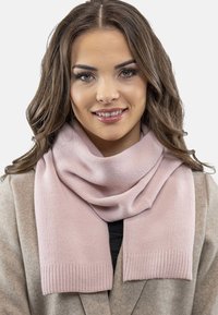 Rosa strikket tørklæde med ribbede ender, hængende løst. Glat tekstur, blødt stof og et varmt udseende, parret med en beige frakke.