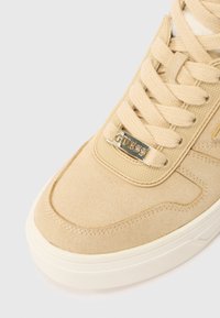 Beżowe zamszowe sneakersy z grubą białą podeszwą, beżowymi sznurowadłami i metalową zawieszką z napisem "GUESS" przy sznurowaniu w okolicy palców.
