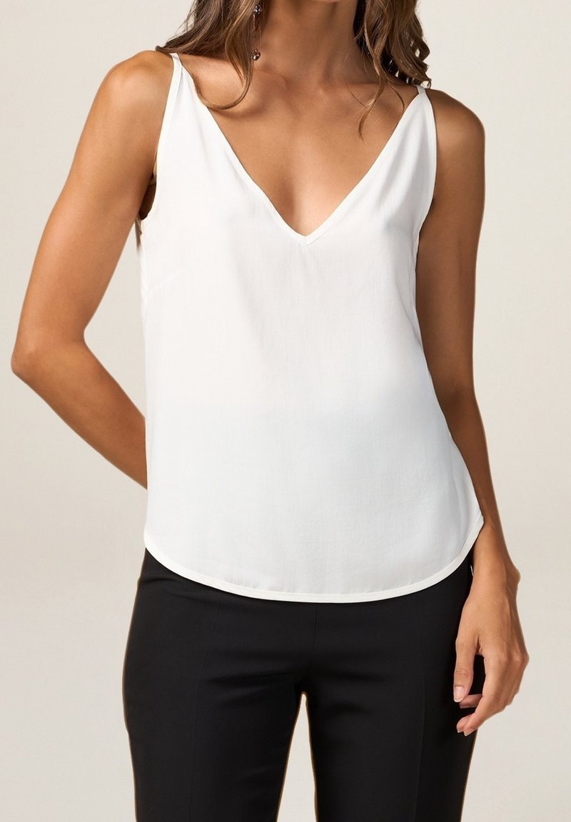 Femme portant un chemisier blanc sans manches à col en V avec de fines bretelles et un pantalon noir, debout devant un fond clair uni.