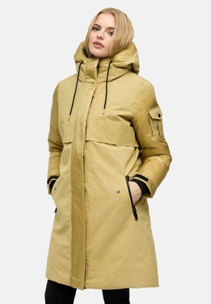Donna con capelli biondi che indossa un lungo cappotto invernale beige con cappuccio e cordini neri, mani nelle tasche con cerniera, su uno sfondo semplice.