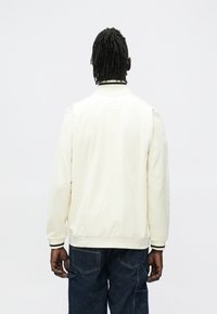 Lysebeige bomberjakke med ribbestrikket krave og manchetter, med tekstureret stof og mørkegrønne accenter, båret over mørke denimbukser.