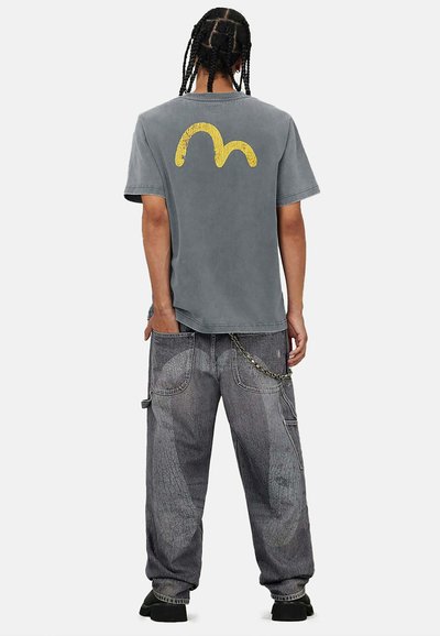 T-shirt grigia in cotone con grafica curva gialla sul retro, abbinata a pantaloni cargo grigi larghi con dettagli testurizzati e catena.