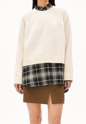 Kvinde iført cremefarvet sweater over sort-hvid ternet skjorte med brun nederdel med frontslids, stående mod hvid baggrund.