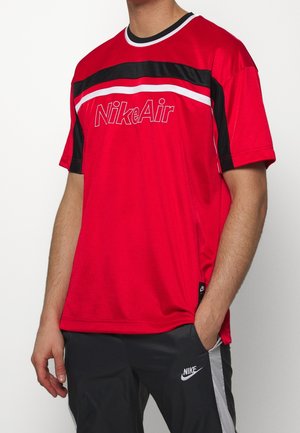 Homme portant un t-shirt rouge Nike Air à manches courtes avec des rayures noires et blanches, et un pantalon de survêtement noir Nike avec une bande latérale blanche.