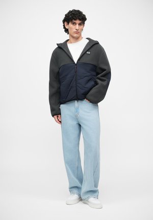Fleece a prošívaná bunda v tmavě šedé a námořnické modři, se zipem vpředu a kapucí. V kombinaci s světle modrými kalhotami s širokými nohavicemi a bílými teniskami.