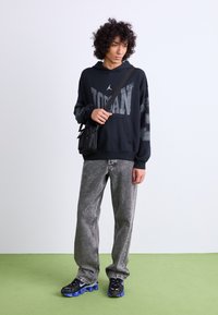 Sort hættebluse med "JORDAN" branding, grå slidte jeans og sorte sneakers med blå accenter. Lille sort taske båret på tværs.