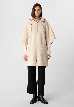 Beige Kapuzenponcho mit einem Frontreißverschluss, gesteppten Taschen und lockerem Schnitt. Kombiniert mit schwarzen Schlaghosen und Stiefeletten.
