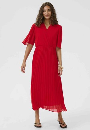 Kobieta z kręconymi włosami, ubrana w czerwoną plisowaną sukienkę midi z krótkimi rękawami oraz czarne sandały na obcasie, stojąca na jednolitym tle.