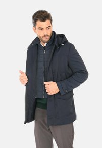 Giacca blu navy con cappuccio, dotata di una fodera interna con zip, due tasche frontali e polsini con bottoni. Tessuto liscio con un design strutturato.