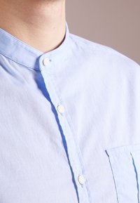 Světle modrá košile s mandarin límcem a bílými knoflíky, odhalující výstřih a horní část hrudníku na neutrálním pozadí.