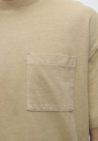 Gros plan d'une chemise beige à manches courtes avec une poche carrée cousue sur la partie gauche de la poitrine.