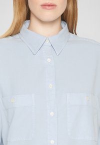 Femme portant une chemise bleu clair à boutons avec deux poches poitrine et des cheveux longs visibles sur les épaules.
