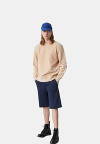 Felpa beige con scollatura tonda, pantaloni corti blu e stivali neri con lacci. Cappellino blu con scritta sulla parte anteriore.