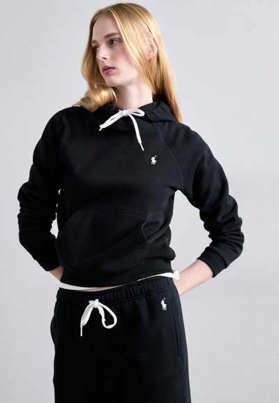 Blondína v čiernej mikine s kapucňou a teplákoch s bielymi šnúrkami a malým bielym logom, stojaca s rukami na bokoch na šedom pozadí.