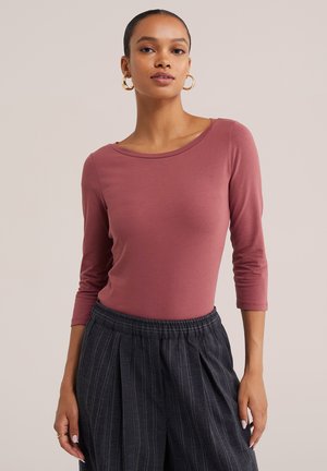 Femme portant des boucles d'oreilles créoles, un haut mauve à manches longues et un pantalon taille haute gris foncé à fines rayures, debout devant un fond uni.