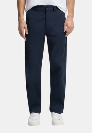 Mand iført marineblå bukser med lige ben, knap og lynlås, kombineret med hvide sneakers og en hvid skjorte, der er stoppet ind.