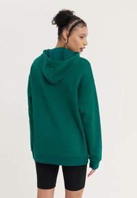 Zelená mikina s kapucí vyrobená z měkké tkaniny s volným střihem. Obsahuje klokánkovou kapsu, ribbed manžety a jednoduchý design na zadní straně.