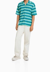 Kortärmad skjorta med vertikala blå och gröna ränder, knäppning fram, tillverkad av lättviktig tyg; tillsammans med elfenbensfärgade byxor och beige sneakers.
