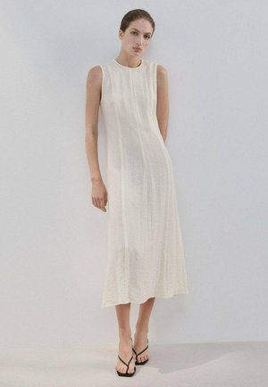 Frau trägt ein ärmelloses, cremeweißes Midikleid mit dezenten vertikalen Nähten, steht vor einem schlichten hellgrauen Hintergrund.