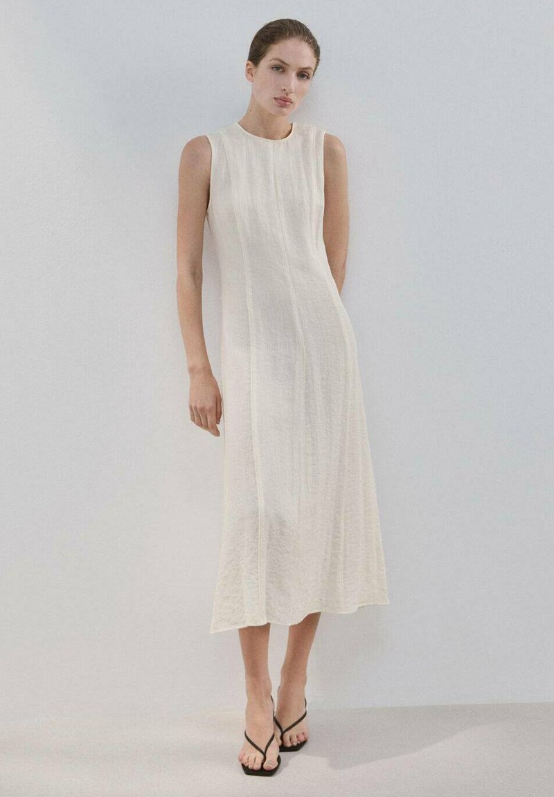 Femme portant une robe midi sans manches de couleur blanc cassé avec des coutures verticales subtiles, debout devant un fond uni gris clair.