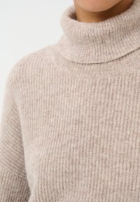 Maglione collo alto a coste color beige realizzato con tessuto morbido, caratterizzato da un colletto alto e una consistenza sottile. In primo piano si vede chiaramente il motivo a maglia.