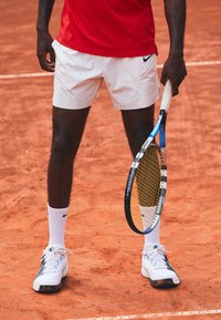 Maglietta atletica rossa, pantaloni corti bianchi, scarpe da tennis bianche con accenti grigi, calze bianche, mentre tiene una racchetta da tennis nera e gialla su un campo in terra battuta.