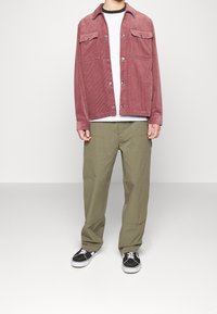 Corduroy jas in stofroze, met twee borstzakken, zilveren knopen; gecombineerd met olijfgroene broek en zwarte sneakers met witte zolen.