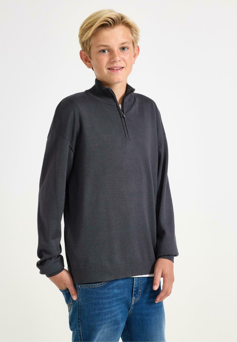 Maglione grigio scuro con zip e collo alto, maniche lunghe, tessitura spessa e vestibilità rilassata, abbinato a jeans blu.