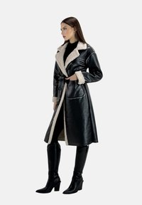 Cappotto in ecopelle nera con collo in pelliccia crema, maniche lunghe, vita segnato da una cintura e tasche laterali, indossato sopra un outfit nero e stivali al ginocchio.