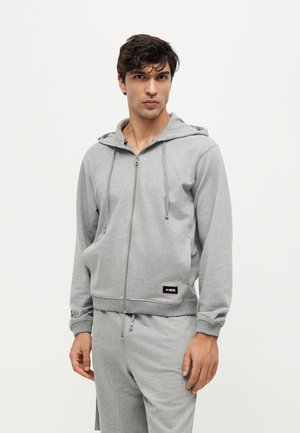 Jeune homme portant un sweat à capuche gris zippé et un pantalon de survêtement assorti, debout sur un fond blanc uni.